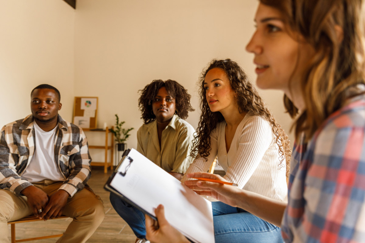 Selective focus shot of young female therapist sitting in circle and holding a clipboard to take notes during a group therapy session.